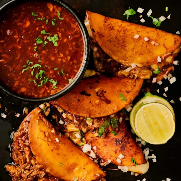 Three Fuego Birria tacos with a side of consommé, garnished with lime and diced onions.