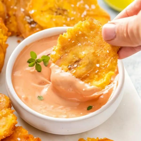 A hand dips a crispy, salted plantain chip into a bowl of creamy orange sauce with herbs.
