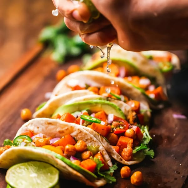A hand squeezes lime juice over a row of Fuego Birria tacos filled with vegetables and chickpeas.